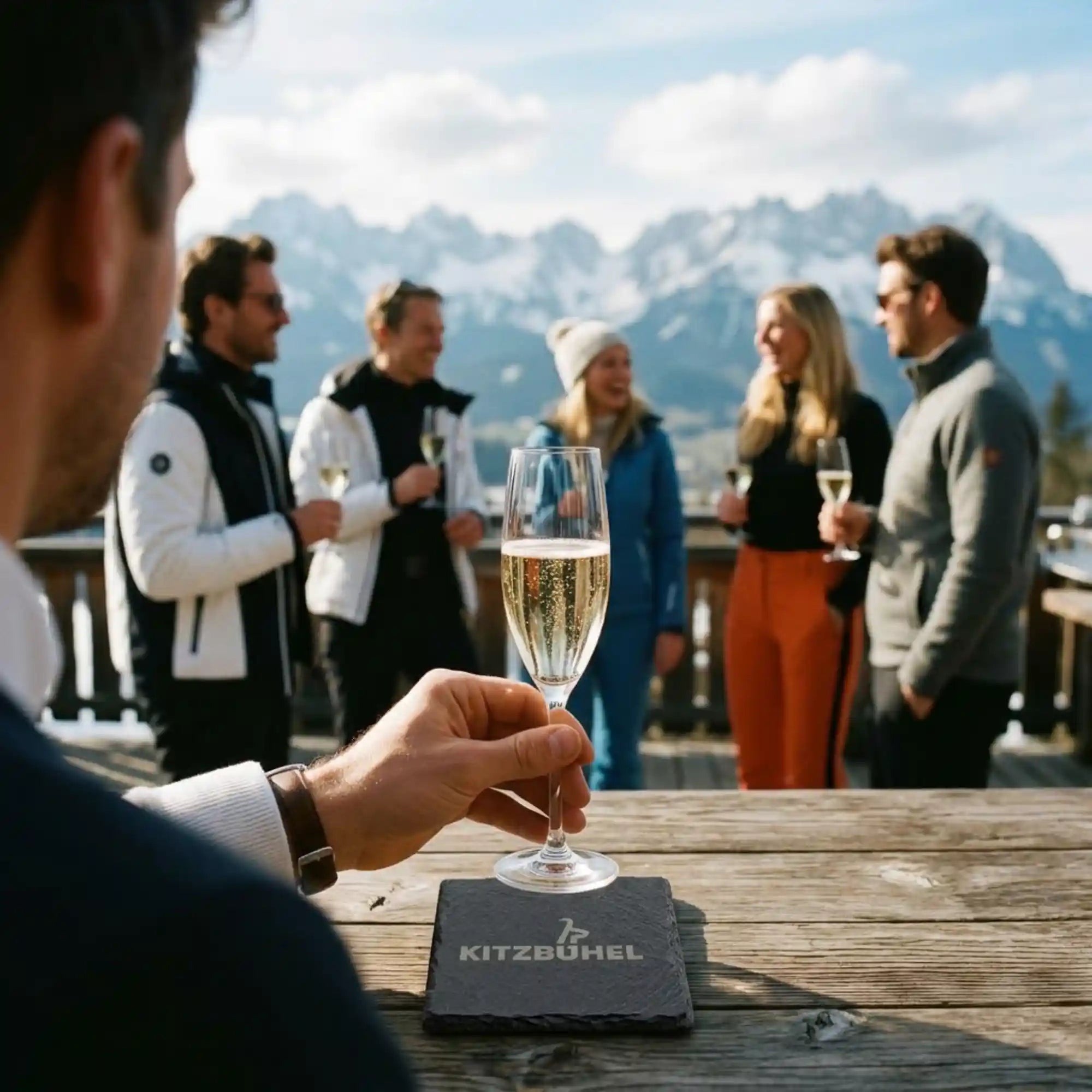 Gravierter Schiefer-Untersetzer mit Kitzbuehel-Schriftzug, auf dem eine Hand ein Champagnerglas abstellt – Terrasse mit Bergblick. Gravierter Schiefer-Untersetzer mit Kitzbuehel-Schriftzug, auf dem eine Hand ein Champagnerglas abstellt – Terrasse mit Bergblick.