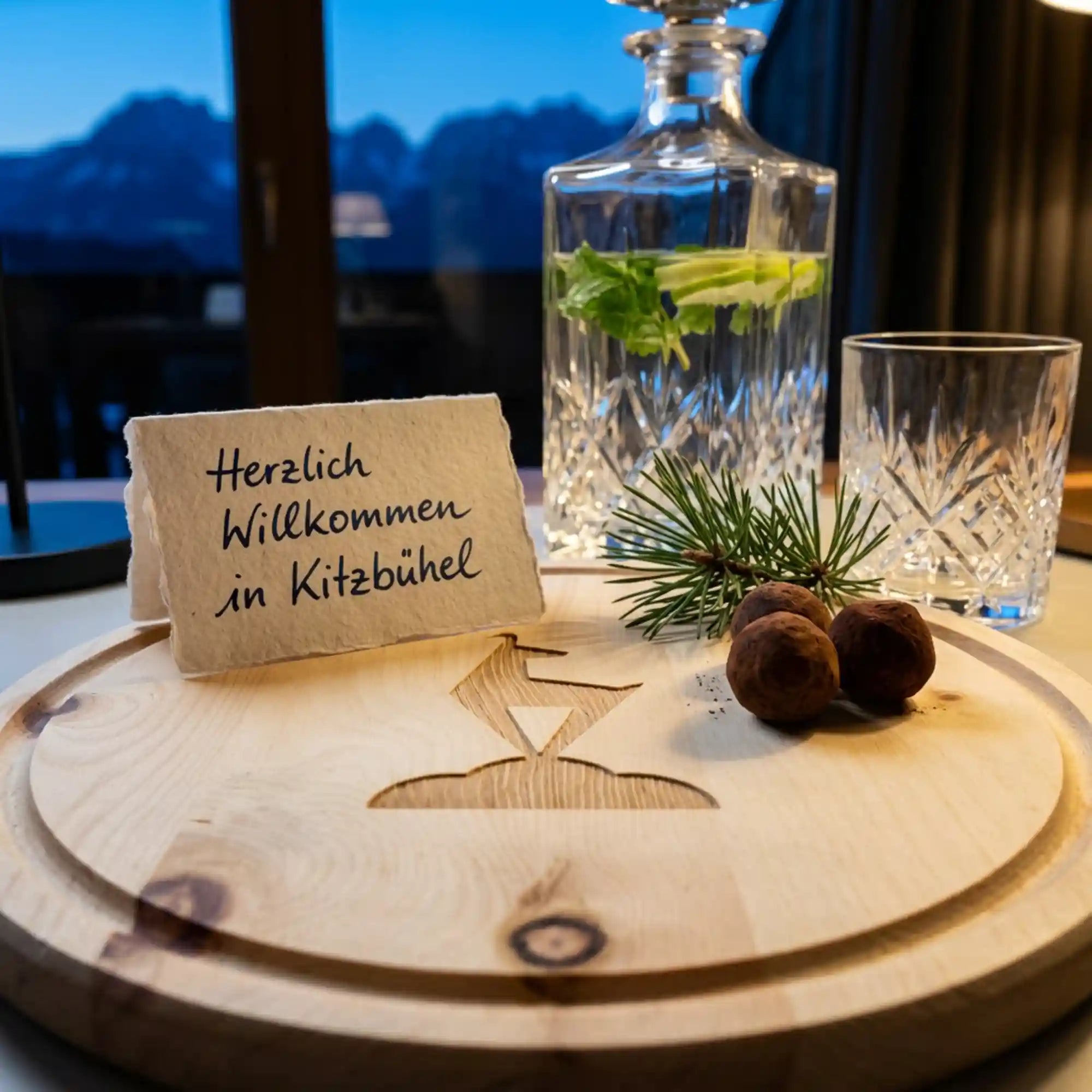 Zirbenholz‑Jausenbrett mit Kitzbuehel‑Logo, Pralinen, Tannenzweig und Grusskarte in einer Hotel‑Suite mit Blick auf den Wilden Kaiser. Zirbenholz‑Jausenbrett mit Kitzbuehel‑Logo, Pralinen, Tannenzweig und Grusskarte in einer Hotel‑Suite mit Blick auf den Wilden Kaiser.