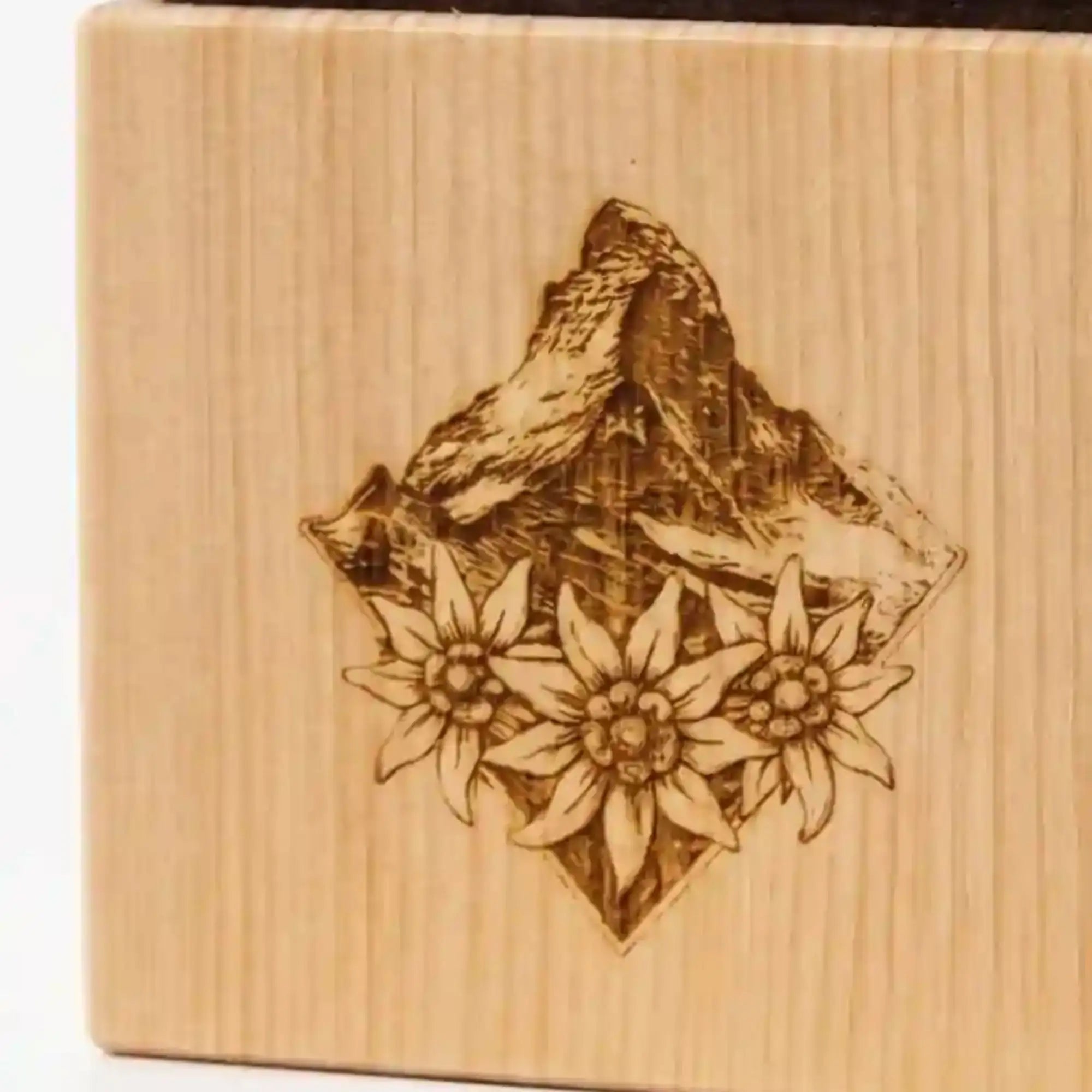 Detailansicht der praezisen Lasergravur mit Alpengipfel und Edelweiß-Blueten auf der gemaserten Holzoberflaeche des Zirbenquaders Alpenblick.