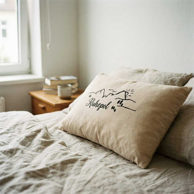Zirbenkissen Zirbenprodukt 30x20 cm mit natürlichen Zirbenflocken und der Aufschrift Ruhepol mit minimalistischer grafik bestehnd aus berg sonne tannenbäumen und wandereren– ohne deko Zirbenprdukt liegt auf dem Bett, wird von Sonnenstrahlen erhellt, Helle bettdecke
