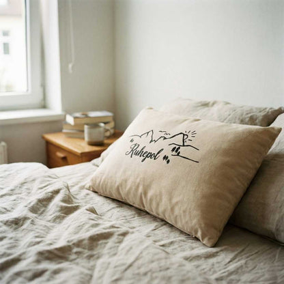 Zirbenkissen Zirbenprodukt 30x20 cm mit natürlichen Zirbenflocken und der Aufschrift Ruhepol mit minimalistischer grafik bestehnd aus berg sonne tannenbäumen und wandereren– ohne deko Zirbenprdukt liegt auf dem Bett, wird von Sonnenstrahlen erhellt, Helle bettdecke