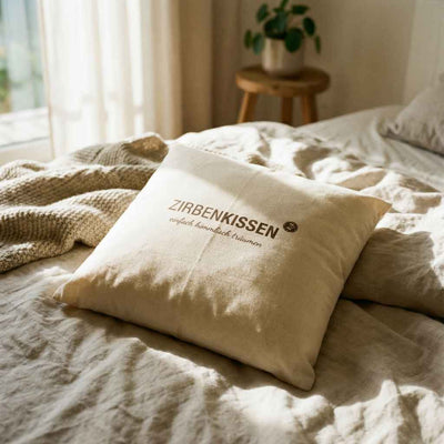 Zirbenprodukt ZirbenKissen 40x40cm “einfach himmlisch träumen” Zirbenprdukt liegt auf dem Bett, wird von Sonnenstrahlen erhellt, Helle bettdecke im Hintergrund pflanze