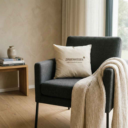 Zirbenkissen ZirbenproduktZirbenprodukt ZirbenKissen 40x40cm “einfach himmlisch träumen” auf grauen sessel daneben eine beige gestrickte decke im hintergrund fenster und ein kleiner tisch mit büchern und einer vase