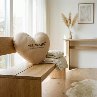 Zirben-Herzkissen beige natur Zirbenprodukt 40x40 cm mit natürlichen Zirbenflocken – handgefertigt in Tirol in einem Großen Lichtdurchfluteten Wohnzimmer auf einer Holzbank neben einer Beigen gestrickten Decke