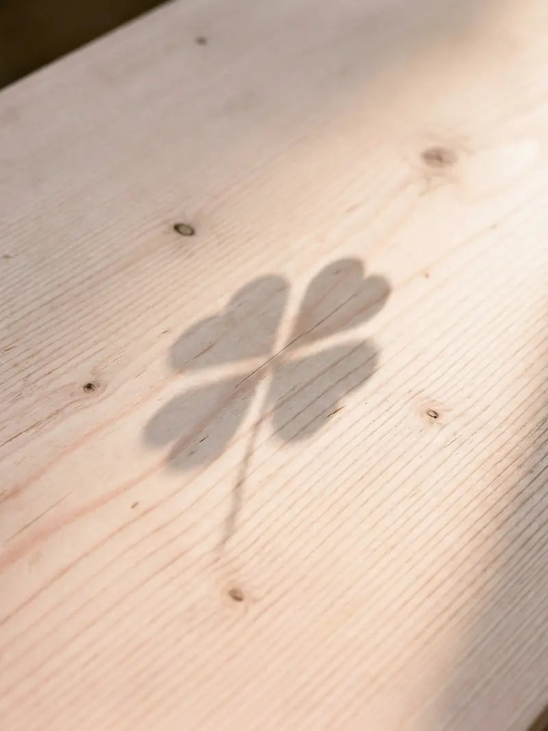 Schatten eines vierblaettrigen Kleeblatts auf Zirbenholz-Tisch bei Sonnenschein: Symbol fuer Glueck und unsere gesamte Auswahl an Fotogeschenken, Zirbe und Wellness in Kitzbuehel.