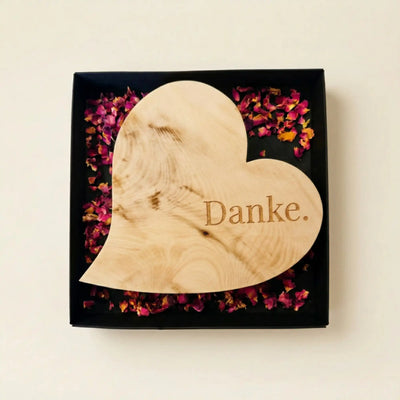 Schlichtes Zirbenholzherz mit der Gravur Danke in einer offenen schwarzen Geschenkbox mit Rosenblueten als Muttertagsgeschenk.