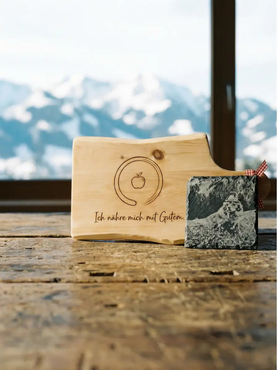 Gegenueberstellung von Fotogravur auf Pinus cembra und Symbolgravur auf Naturschiefer vor einem alpinen Bergpanorama - Manufaktur bei Kitz.