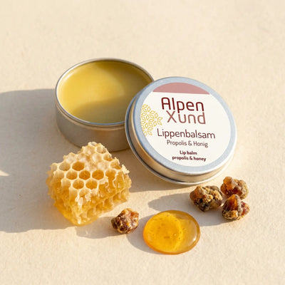 Offene silberne Lippenbalsam Dose mit Honigwabe, Honigtropfen und Propolis Harz auf beigem Papier