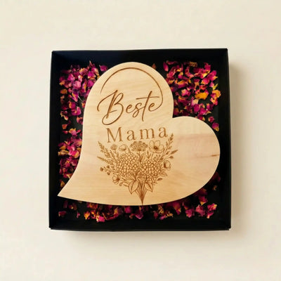 Zirbenholzherz mit Blumenstrauss-Gravur und Schriftzug Beste Mama in einer offenen schwarzen Geschenkbox mit Rosenblueten zum Muttertag.