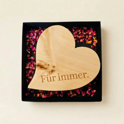 Zirbenholzherz mit der Gravur Fuer immer in einer offenen schwarzen Geschenkbox mit kleinen Rosenblueten als Dankeschoen zum Muttertag.