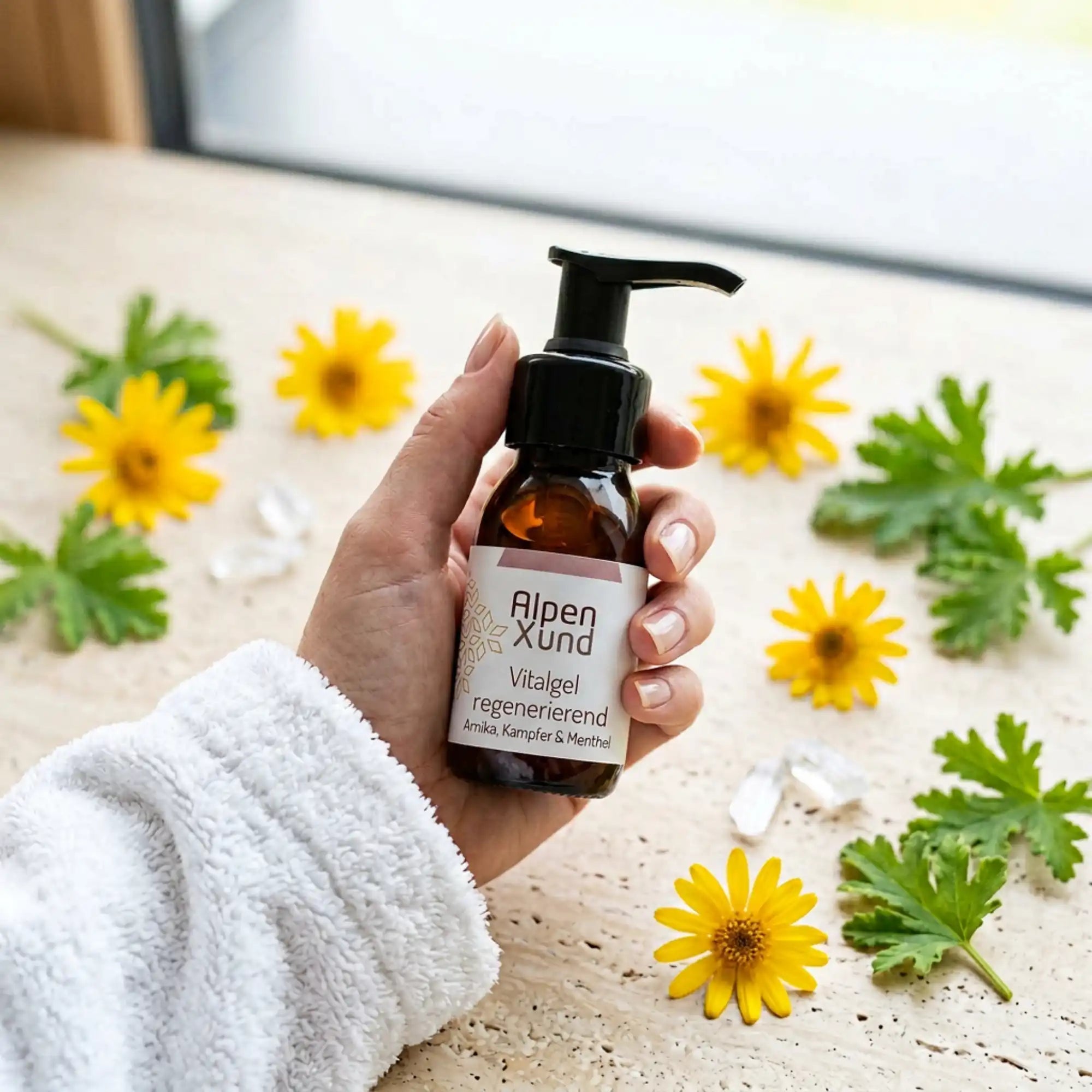 Eine Hand im weissen Bademantel haelt eine Vitalgel mit Arinka. Hintergrund mit gelben Arnika Blueten, Citronella und Menthol Kristallen auf Beige.