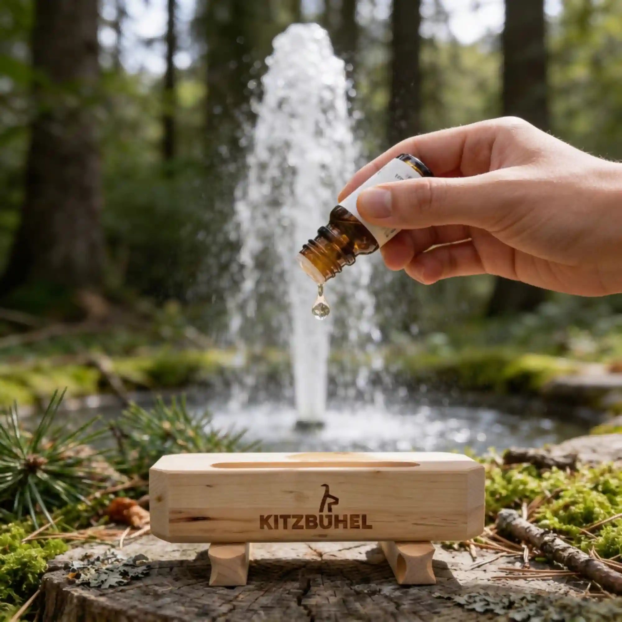 Hand traeufelt Zirbenoel in den Zirbenduft‑Brunnen Kitz‑Edition, im Hintergrund Natur und ein Wasserbrunnen mit Fontaene.