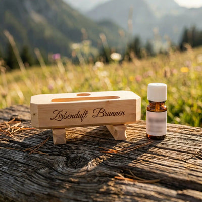 Zirbenduft-Brunnen und 10ml Zirbenoel Flasche nebeneinander auf rustikalem Altholz vor sonniger Bergwiese.