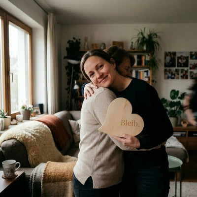 Zirbenherz mit Gravur „Bleib.“ als emotionales Muttertagsgeschenk – zwei Frauen umarmen sich herzlich in einem gemuetlichen Wohnzimmer.