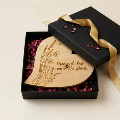 Geoeffnete schwarze Geschenkbox zum Muttertag 2026 mit Zirbenherz Mama. du bist unser Herzstueck auf duftenden Rosenblueten.